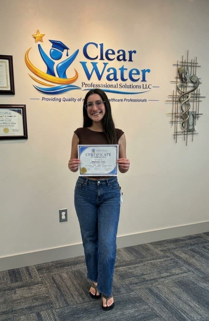 Clear Water graduate smiling with a certificate of completion at the studio logo wall